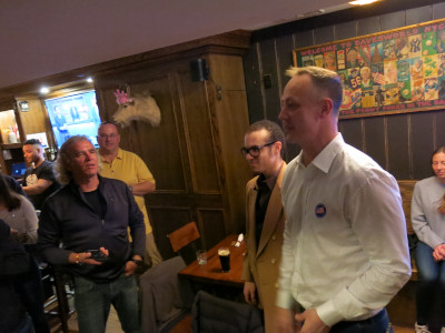 Photo of a man in a white shirt speaking to a crowd in a bar.