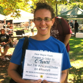 Photo of a woman holding up a sign that says: I want to bike to C-Town but I'm #2scared2bike on Route 9.