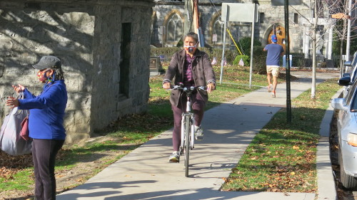 Woman cycling on the sidealk towards us. She's wearing a purple coat, shirt and pants.
