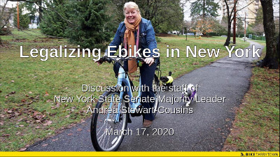 Woman riding an ebike on a paved path with two little dogs behind her.