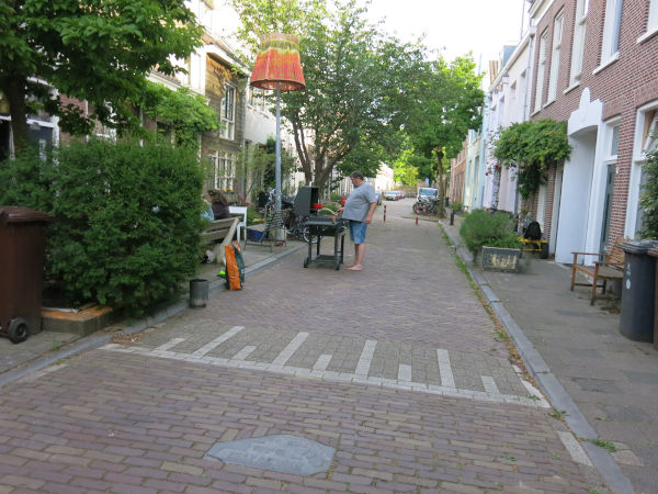 Photo of a person standing on the side of a small street, grilling food for their family sitting in chairs on the sidewalk. Art is on the light pole