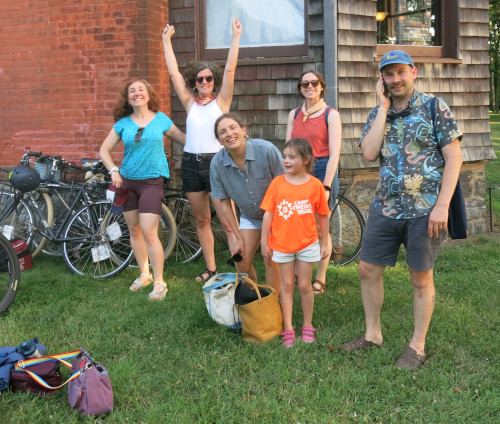 Photo of 4 adults and a kid happily standing next to their bikes on the grass next to a building.