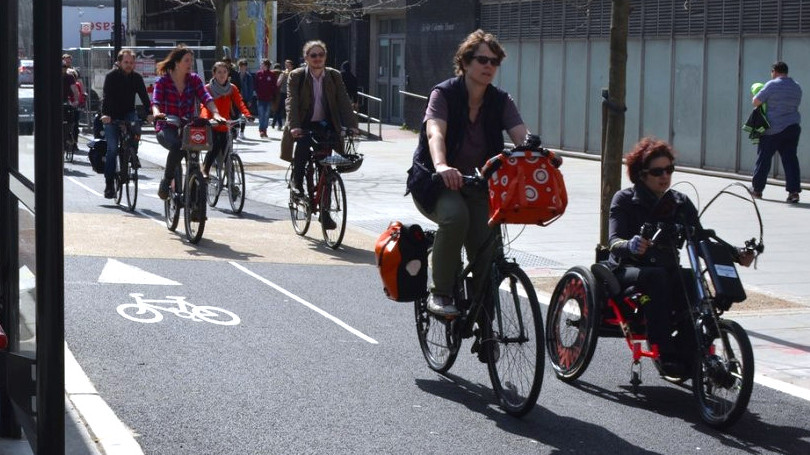 Photo of people using a 2-way protected cycling lane. One of the people is in a wheelchair with a clip on hand cycle attachment with electric assist.