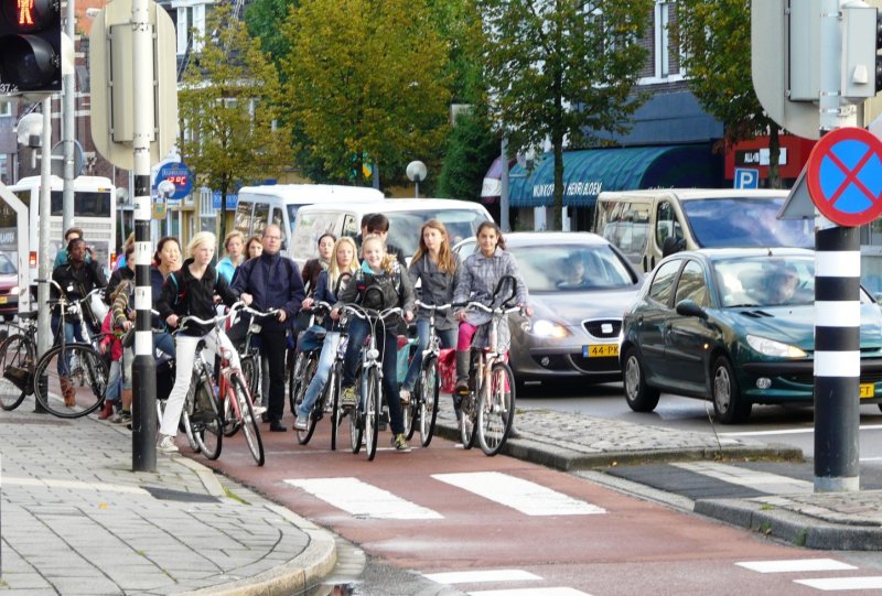 Large group of people (mostly teenage girls) on bicycles waiting at a traffic signal on a protected cycle lane.