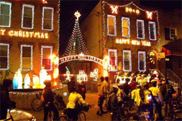 Photo of a group of cyclists standing in front of two buildings decorated with christmas lights.